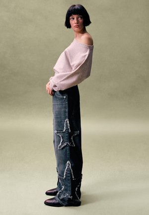 Nadmiernie duży jasnoróżowy sweter z odkrytymi ramionami, zestawiony z ciemnoniebieskimi jeansami ozdobionymi postrzępionymi naszywkami w kształcie gwiazd oraz brązowymi botkami za kostkę.