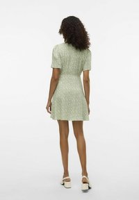 Robe florale verte avec des manches courtes, taille cintrée, ourlet évasé et tissu lisse et léger. Le mannequin porte des sandales à talons carrés de couleur crème.