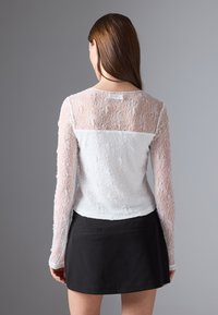 Kvinne med langt brunt hår iført en hvit blond langermet topp og et svart skjørt, stående med ryggen mot kameraet mot en grå bakgrunn.