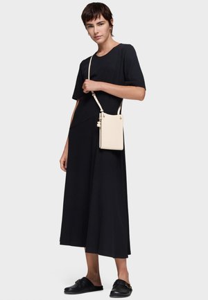 Mujer con un vestido midi negro y zapatos negros sin cordones, sosteniendo un pequeño bolso cruzado crema con un colgante de oso.