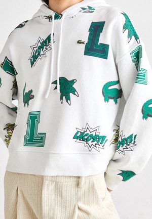 Biała bluza z kapturem z zielonymi wzorami krokodyla, pazura, litery L i logo Lacoste, noszona z beżowymi, teksturowanymi spodniami.