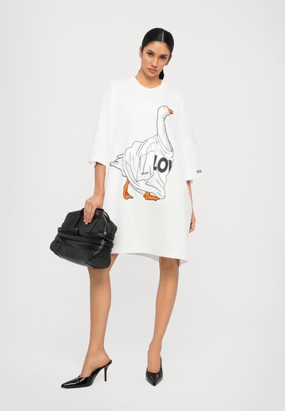 Robe oversize blanche avec un motif de oie, sac à main noir et escarpins noirs brillants à bout pointu au design épuré.