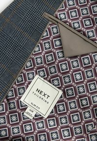 Tissu texturé gris avec un motif à carreaux bleu, associé à une doublure bordeaux ornée de motifs géométriques blancs ainsi qu'une étiquette de soin.