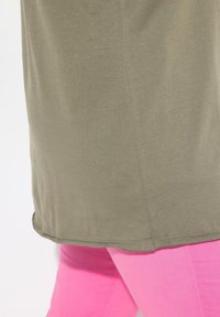 Chemise vert olive avec une texture douce et un ourlet arrondi, associée à un pantalon rose taille haute. La chemise présente des détails de couture subtils.