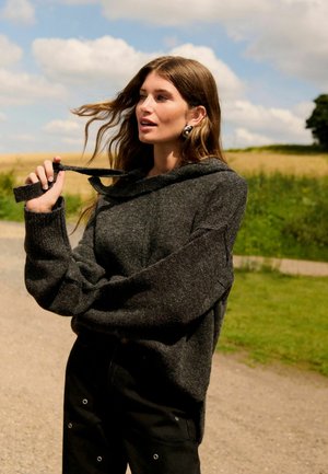 Dark gray knitted sweater with a hood, featuring oversized sleeves and a tied drawstring. Worn with black pants adorned with grommets.