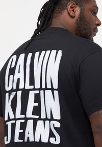 Černé tričko s velkým bílým nápisem "CALVIN KLEIN JEANS." Tričko má kulatý výstřih a krátké rukávy.
