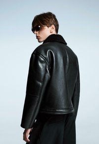 Crna kožna jakna s krznenim ovratnikom, oversized kroj, izraženi rameni šavni detalji, uparena s tamnim hlačama koje su nosene naopako.