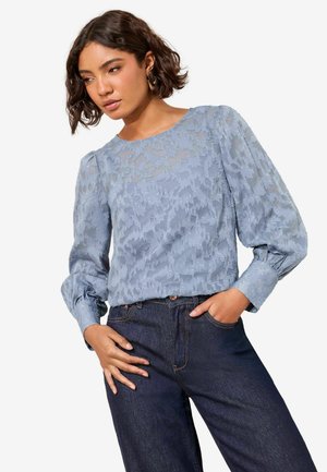 Világoskék blúz texturált virágmintával, kerek nyakkal és fodros ujjakkal. Sötét farmernadrággal párosítva. Az anyag lágynak és áttetszőnek tűnik.