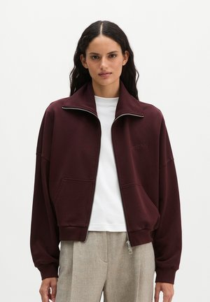 Vrouw met lang donker haar die een open donker bordeauxkleurige jas met ritssluiting draagt over een wit overhemd en lichtbeige broek, staand tegen een effen achtergrond.