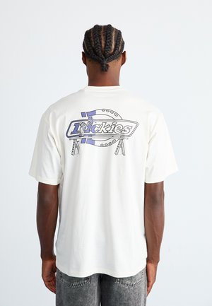 Man with braided hair wearing a white T-shirt with a blue and black "Dickies" logo and illustration on the back, paired with gray jeans.