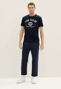Marineblå bomull t-skjorte med "TOM TAILOR" grafikk, sammen med mørk marineblå bukser. Modell iført hvite sneakers, avslappet passform.