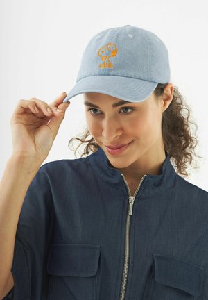 Femme aux cheveux bouclés portant une veste bleue zippée et une casquette bleu clair avec une broderie orange de Snoopy, ajustant la casquette de la main droite.