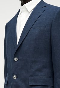 Άνδρας που φοράει μπλε σκούρο σακάκι με υφή πάνω από ένα λευκό πουκάμισο με κουμπιά, εμφανίζοντας το πάνω μέρος του κορμού και τις λεπτομέρειες του πέτου.