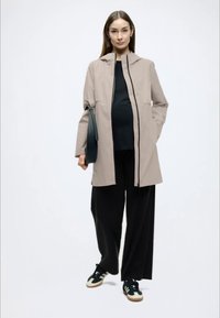 Μπεζ αδιάβροχο μπουφάν με ψηλό γιακά, μαύρο εσωτερικό τοπ, φαρδιά μαύρη παντελόνα και σκουρόχρωμα αθλητικά παπούτσια. Διαθέτει φερμουάρ και πλαϊνές τσέπες.