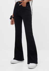 Schwarze, aus Denim gefertigte, weit geschnittene Jeans mit hoher Taille, Vordertaschen und dezenten Nähdetails. Kombiniert mit weißen Sneakern.