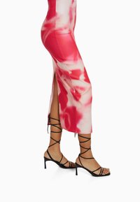 Robe ajustée rose et blanche à motifs tie-dye avec fentes sur les côtés, assortie à des sandales à talons noires avec des lanières croisées et des orteils ouverts.