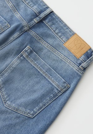 Vue arrière d'un jean en denim bleu clair montrant la poche arrière droite, la passoire de ceinture, les détails de couture, et une étiquette de marque en cuir marron clair sur la ceinture.