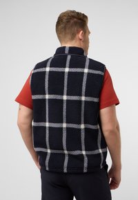 Navy Fleece-Weste mit einem weißen und grauen Karomuster. Hoher Kragen und ärmelloses Design. Getragen über einem roten kurzärmeligen Hemd.