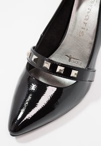 Chaussure noire en cuir verni à bout pointu avec une bride sur le dessus ornée de clous carrés argentés et une semelle intérieure argentée texturée.