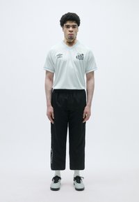 Gaišzils futbola krekls ar melnbaltiem svītrainiem akcentiem, apvienots ar melnām biksēm un gaiši ziliem sporta apaviem, uz baltā fona.