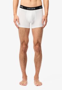 Fehér boxer alsónadrág rugalmas anyagból, fekete Lacoste logós derékpánttal; testhezálló formájú és sima tapintású.