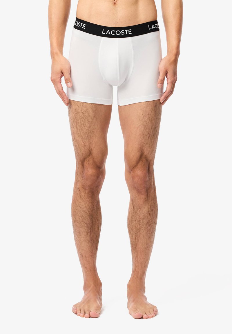 Culotte boxer blanche en tissu extensible avec une ceinture noir arborant le logo Lacoste ; elle présente une coupe ajustée et une texture douce.