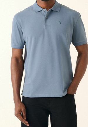 Homme portant un polo bleu clair à manches courtes avec un petit logo foncé sur la poitrine, associé à un pantalon noir.