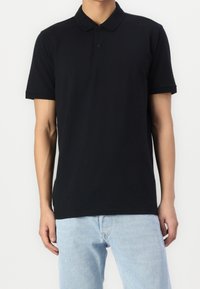 Man som bär en enfärgad svart kortärmad pikétröja som kombineras med ljusblå jeans mot en vit bakgrund.