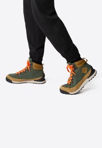 Buty trekkingowe z zielonym materiałowym cholewką, brązowymi zamszowymi akcentami, pomarańczowymi sznurowadłami, czarną gumową podeszwą i wspierającym kołnierzem.