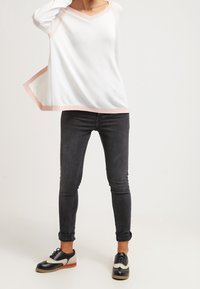 Persona che indossa una maglietta bianca a maniche lunghe con bordi rosa, jeans skinny scuri arrotolati alla caviglia e scarpe oxford bianco e nere con suole rosse.