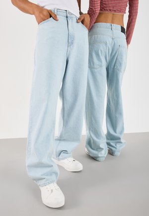 Deux personnes portant des jeans bleu clair à jambes larges et des baskets blanches, l'une face avant avec les mains dans les poches, l'autre face arrière.