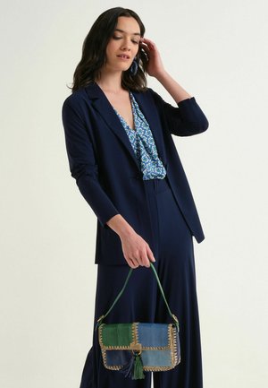 Donna con blazer blu navy e pantaloni che tiene una borsa patchwork blu-verde, indossa una camicetta blu fantasia e orecchini abbinati.