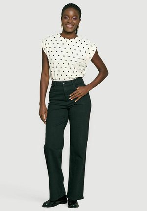 Vrouw staand en lachend, draagt een wit mouwloos polkadot topje, zwarte wijde hoge taille broek en zwarte schoenen.