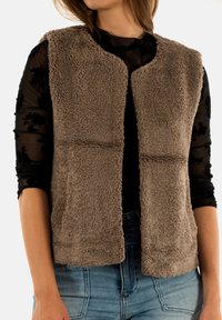 Donna che indossa un gilet senza maniche marrone e peloso sopra una blusa nera trasparente con fiori, e jeans blu, con le braccia rilassate ai lati.