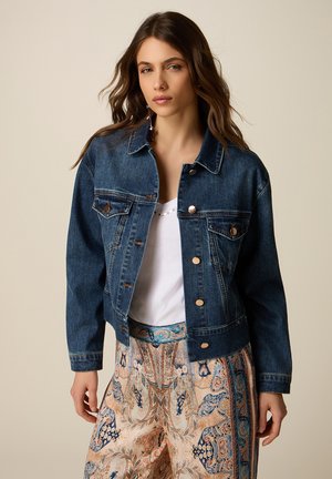 Donna che indossa una giacca di jeans blu sopra una maglietta bianca, abbinata a pantaloni beige a zampa con motivo, su uno sfondo beige uniforme.