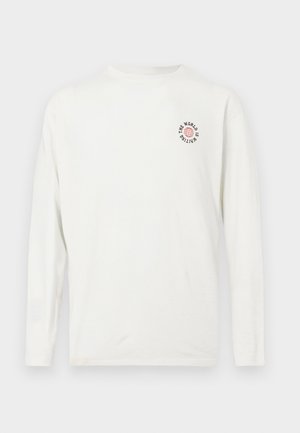 Witte lange mouwen shirt met een kleine rode wereldbol grafiek en zwarte tekst "DE WERELD WACHT" op de bovenste linkerkant van de borst.