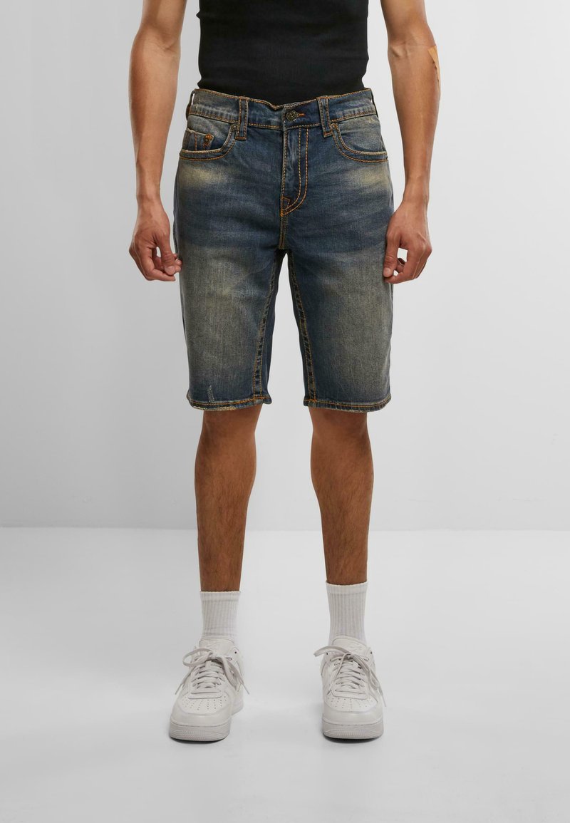 Muž v bledých džínsových kraťasoch po kolená, bielych ponožkách, bielych teniskách a čiernej bezrukávovej blúzke, stojí na jednomfarebnej šedej podlahe.