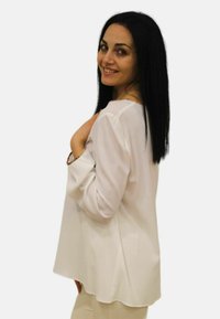 Blusa bianca a maniche lunghe con un fit rilassato, texture liscia e orlo arrotondato. Il modello posa di lato, tenendo la mano vicino alla spalla.