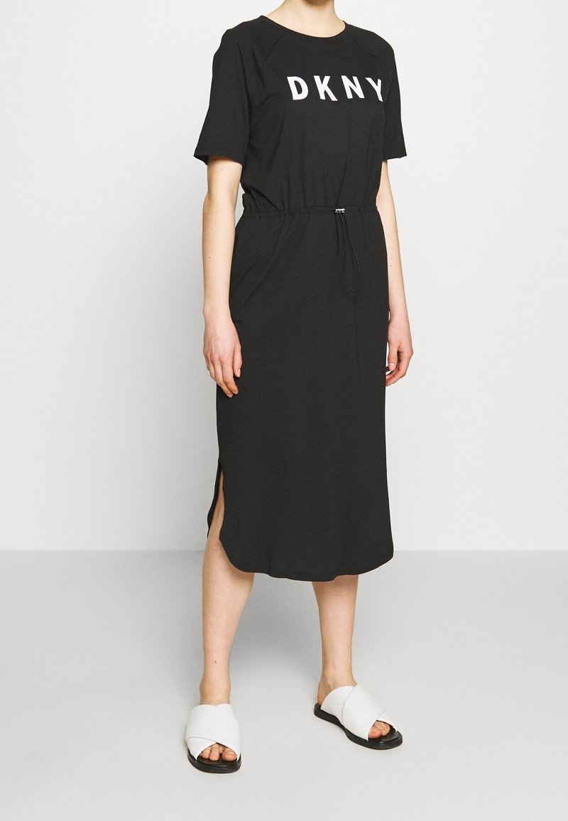 Personne portant une robe midi noire avec le logo DKNY, manches courtes, fentes latérales et sandales blanches à enfiler.