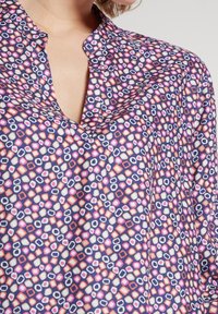 Blouse multicolore avec un motif géométrique composé de formes violettes, roses et blanches. Le tissu a une texture lisse et un col pointu.
