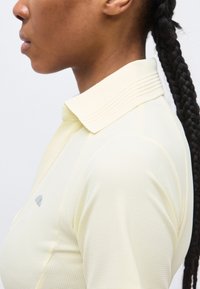 Vue de profil d'une personne aux longs cheveux tressés portant un polo jaune clair texturé avec un col côtelé.