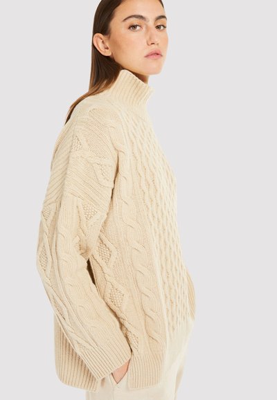 Suéter beige de cuello vuelto de punto, con patrones de trenzado, dobladillo acanalado y hombros caídos, hecho de un material suave y texturizado.