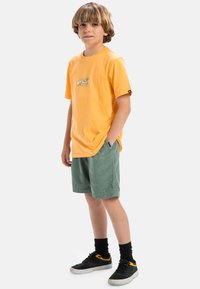 Joven vistiendo una camiseta de algodón amarilla con un gráfico, combinada con pantalones cortos de pana verde y zapatillas negras con acentos amarillos.