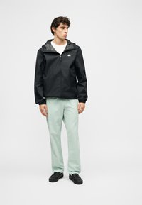 Νεαρός άνδρας που φοράει μαύρο τζάκετ με φερμουάρ και κουκούλα, λευκό πουκάμισο, ανοιχτοπράσινο παντελόνι και μαύρα παπούτσια, στέκεται μπροστά σε μονόχρωμο φόντο.