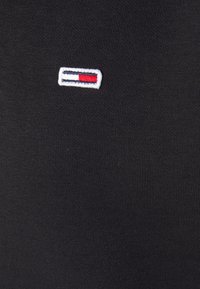 Camiseta de algodón negra con un primer plano de un pequeño logo bordado que presenta franjas rojas, blancas y azules sobre un fondo blanco rectangular.