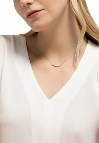 Zilveren ketting met kleine, ronde kralen in een gebogen patroon, gedragen boven een wit V-hals top.