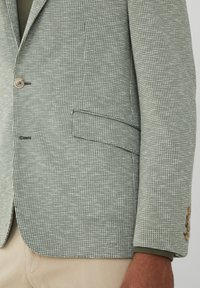 Svetlozelený štruktúrovaný blazer s vzorom houndstooth, obsahujúci dva gombíky, jedno náprsné vrecko a kontrastné švy.