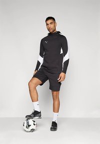 Schwarz-weißes Sportoutfit mit Hoodie und Shorts, kombiniert mit schwarzen Schuhen und weißen Socken, das sich auf einen Fußball lehnt.