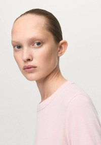 Portret osebe z ravnimi lasmi, ki nosi svetlo roza majico s kratkimi rokavi. Ozadje je nevtralno in gladko.