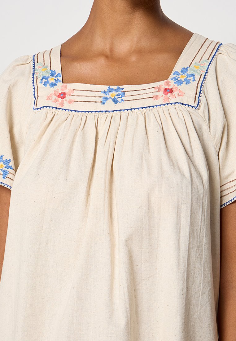 Crème blouse met vierkante halslijn, geborduurd met blauwe en roze bloemen, gedragen door een persoon met mediumbruine huid.